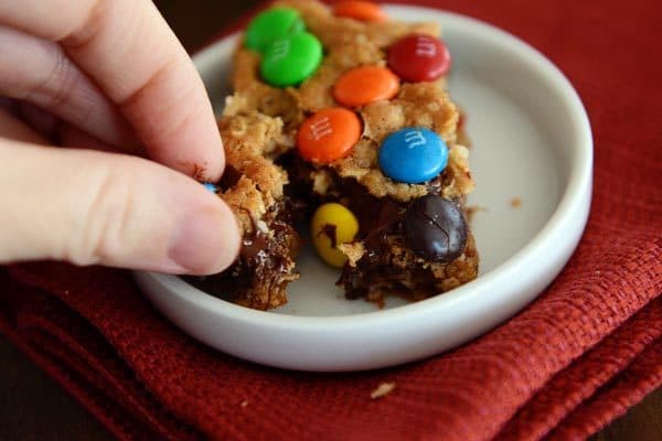 A hand taking a chunk off of an M&M topped oat bar in a small white ramekin.