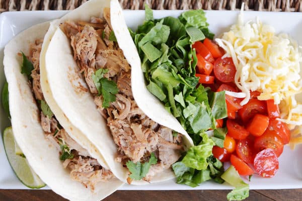 A white tray of pork tacos, salad, cherry tomatoes, and shredded cheese. 
