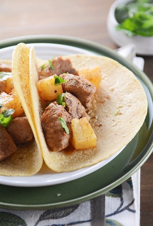 Two pork and pineapple tacos in corn tortillas on a white plate.
