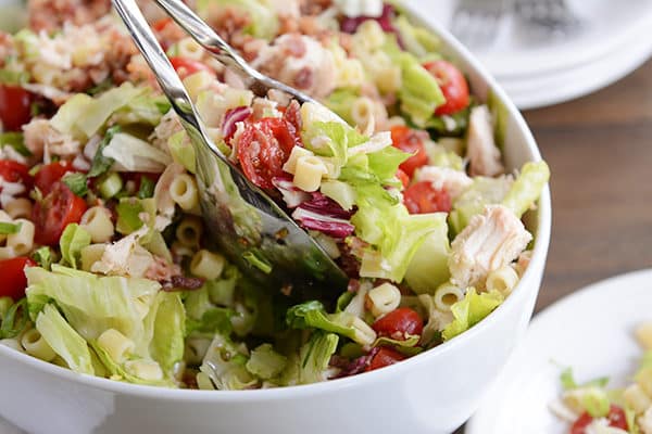 a white bowl of chicken, pasta, and tomato chopped salad.