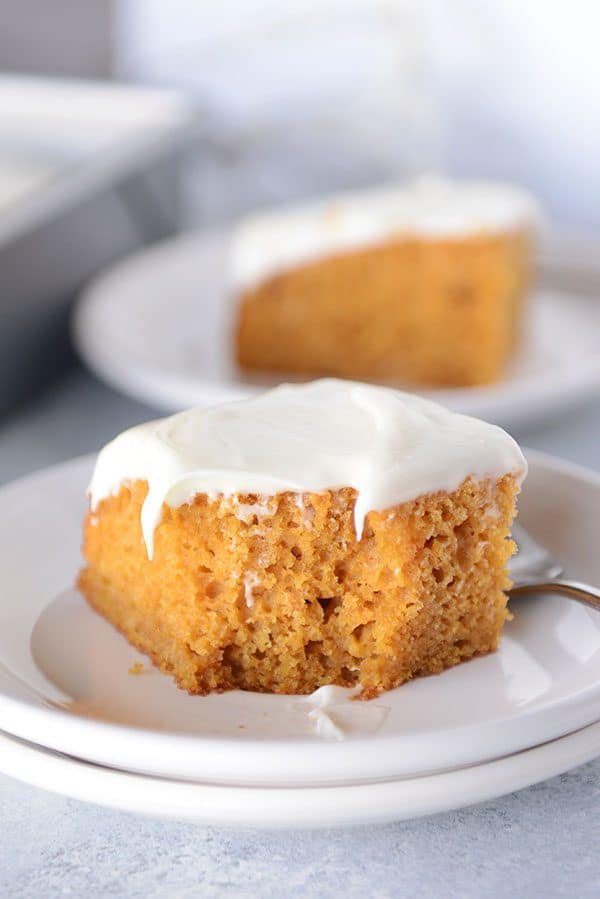 A piece of cream cheese frosted pumpkin cake with a bite taken out on a white plate.