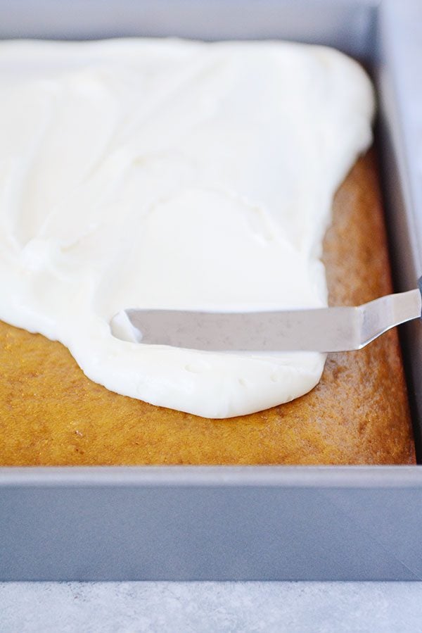 A pan of pumpkin bars getting frosted with a metal spatula.