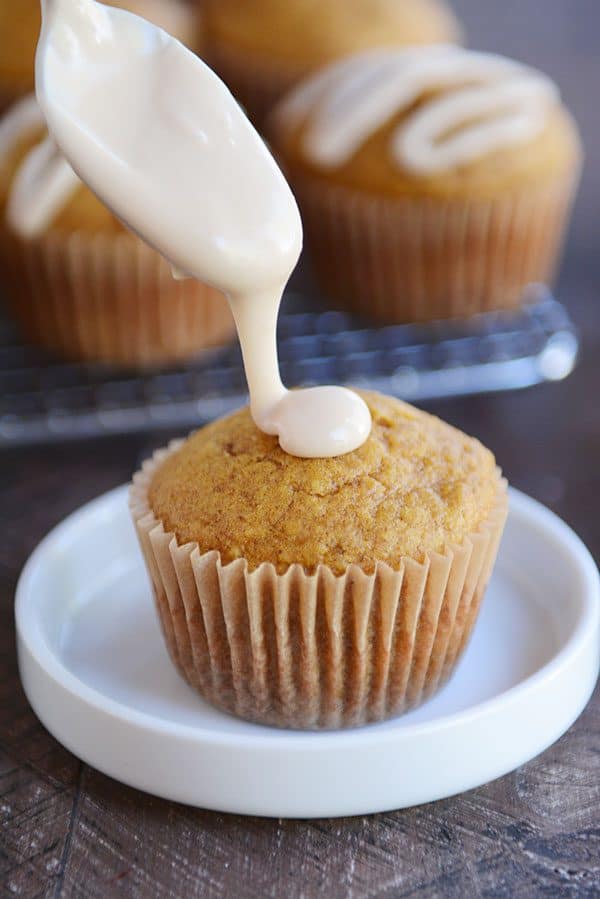 A spoon drizzling white frosting onto a pumpkin muffin.