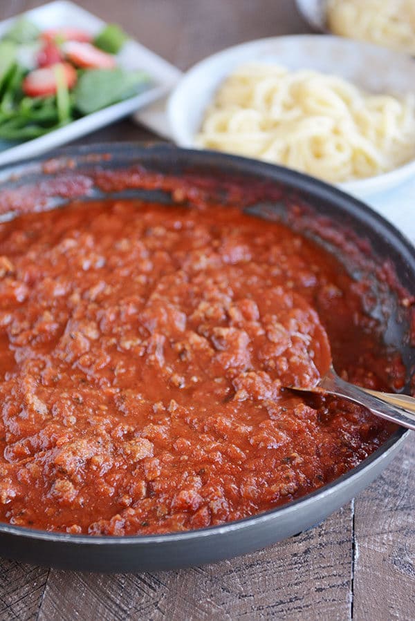 A skillet full of meaty, red spaghetti sauce, with a green salad, and spaghetti noodles in the background.