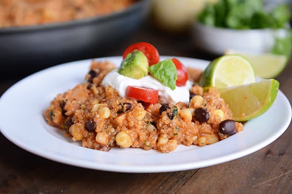 A white plate with a serving of a quinoa enchilada casserole topped with sour cream, avocados, tomatoes, and lime slices.