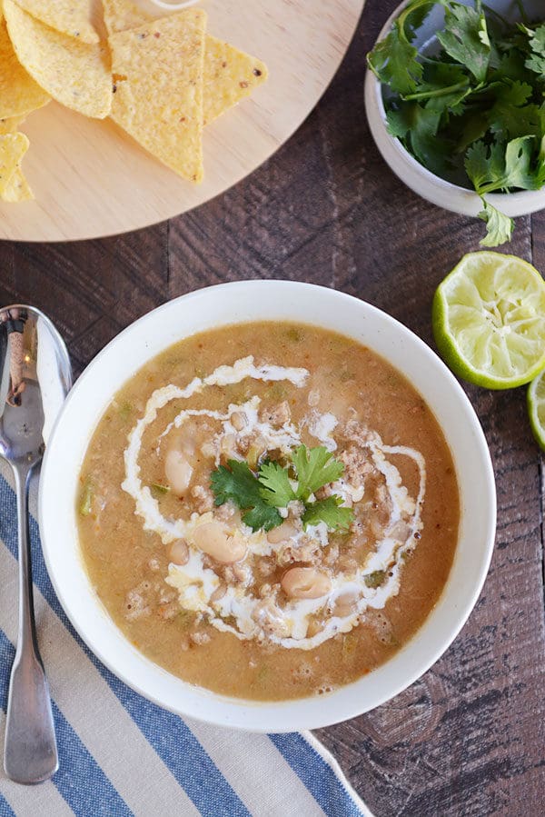 Top view of a bowl of white bean chili with limes, parsley, and tortilla chips beside it.