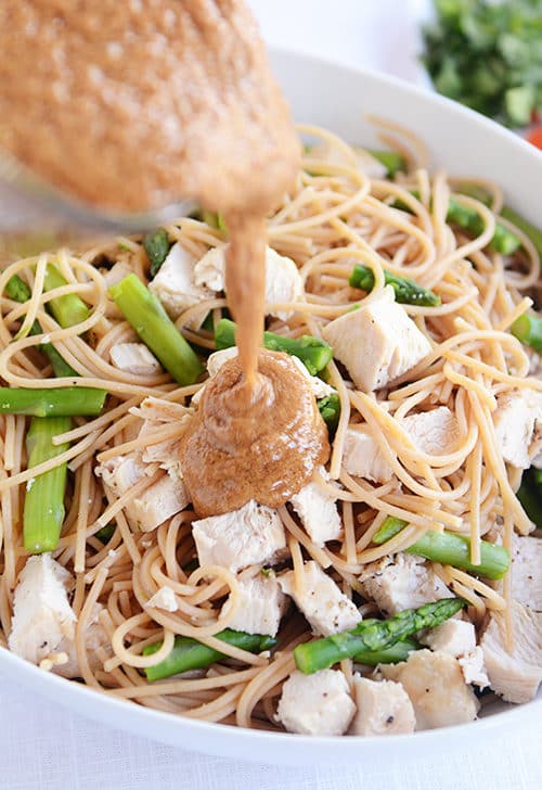 A bowl of cooked pasta, chicken cubes, and asparagus pieces getting sauce poured over the top. 