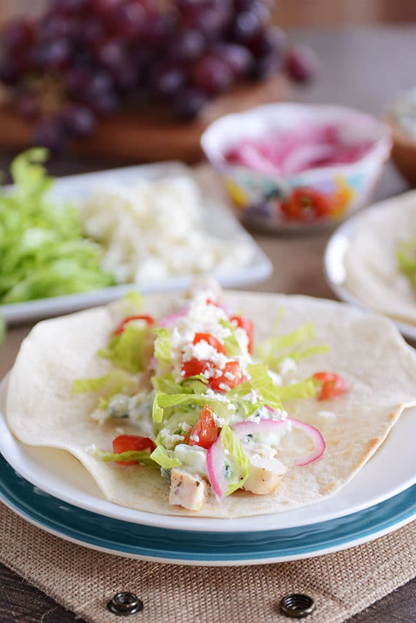 And unwrapped Greek chicken burrito with a platter of toppings in the background.