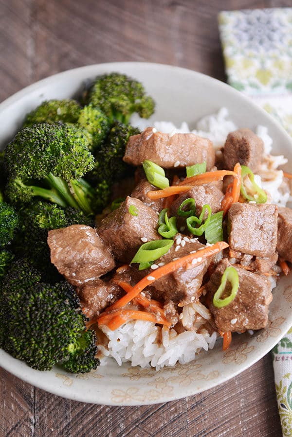 A bowl full of white rice with cubes of beef, shredded carrots, and green onions on top, and broccoli on the side. 