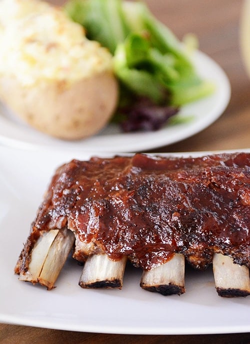 A rack of ribs smothered in sauce on a white platter in front of a plate of potatoes and salad.
