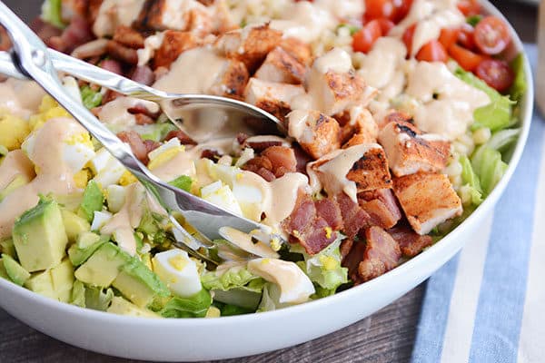 Metal tongs taking a helping of salad out of a white oval bowl full of grilled chicken cobb salad.
