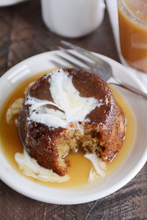 A small brown cake, with a bite taken out, covered with sticky toffee sauce and cream on a white plate.