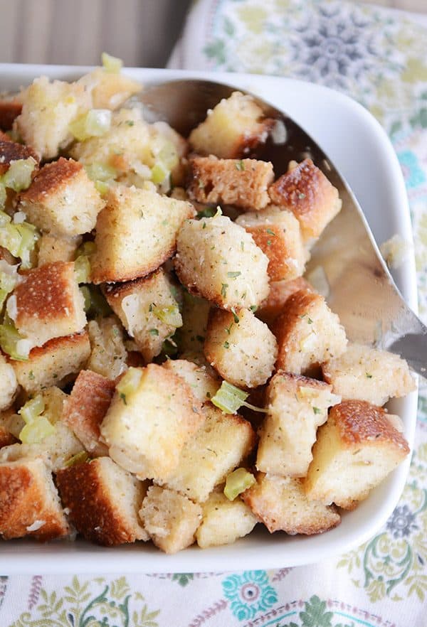 A white tray of stuffing with a large metal spoon taking a scoop out.