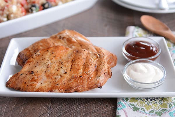 Two grilled chicken breasts next to two small glass cups of dipping sauce on a white platter.