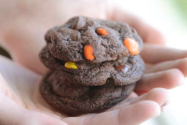 A hand holding three chocolate M&M cookies stacked on top of each other.