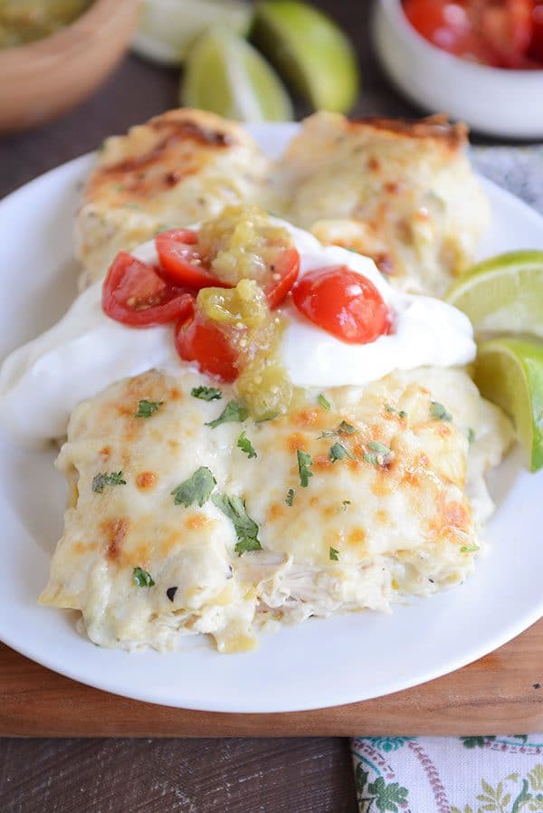 Two baked green chile chicken enchiladas side-by-side on a plate, topped with sour cream and cherry tomatoes and sliced limes on the side.