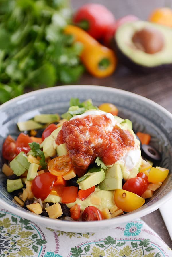 A bowl full of chopped veggies, avocado, tomatoes, cilantro, salsa, and sour cream.