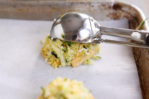 A cookie scoop putting a mound of cheesy uncooked zucchini batter on a cookie sheet.