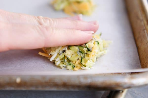 A hand patting down a cheesy uncooked zucchini bite on a cookie sheet.