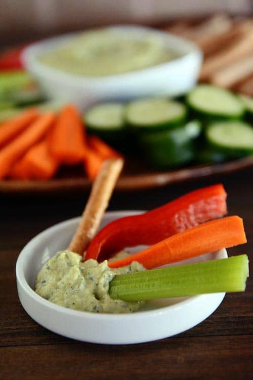 A tray of cut up vegetables and zucchini hummus, and a small white ramekin with hummus and some veggie sticks inside.