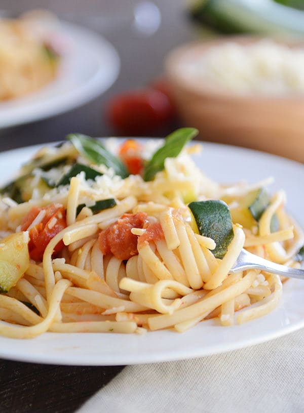 A fork taking a bite of zucchini and tomato pasta off of a white plate.